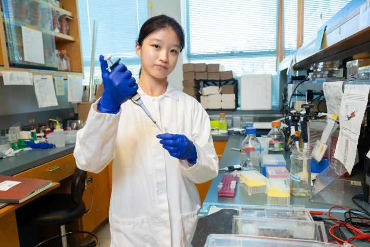 Student wearing gloves and white lab coat works at lab station