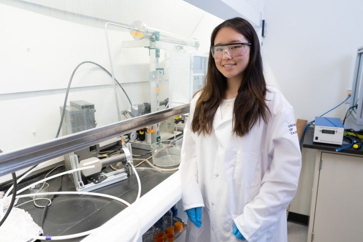 Student wearing gloves and white lab coat poses in front of lab hood