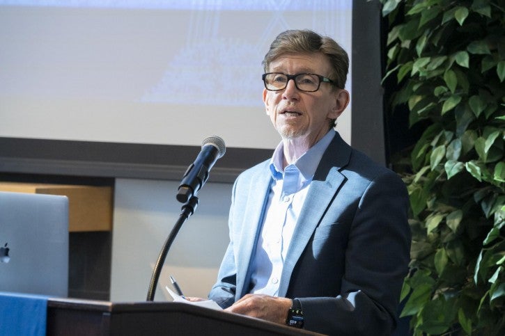 A man wearing glasses and a suit stands at a podium and speaks into a microphone.