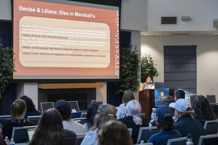 A woman stands at a podium and speaks to an audience sitting in front of her. The presentation screen behind her shows an orange slide with research details on it.