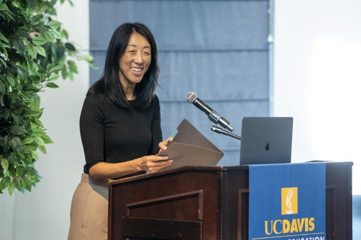 A woman wearing a black shirt and tan pants stands at a podium and smiles. A laptop is open in front of her.