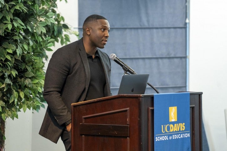 A man wearing a blazer stands at a podium and speaks into a microphone. One of his hands is in his pocket.