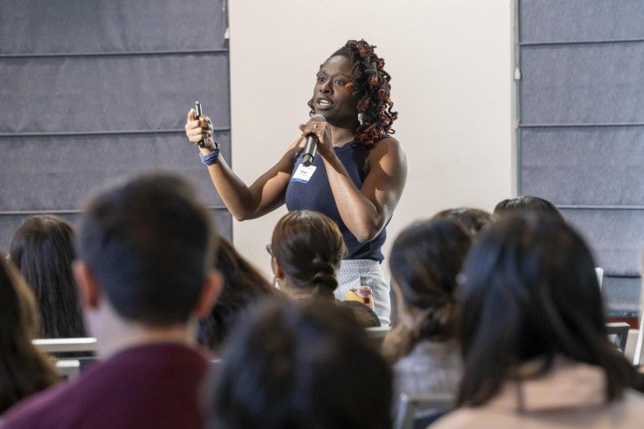 A woman stands in front of a group of people and speaks into a microphone while gesturing with her hand.