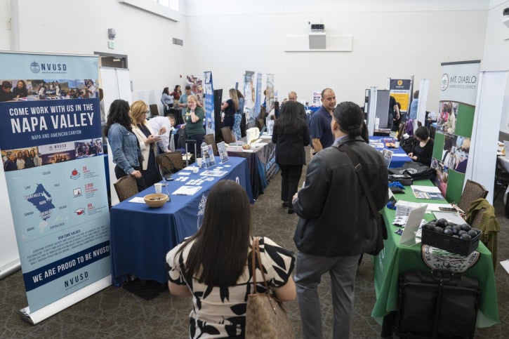 A bustling career fair in a bright room with tables from different organizations. Attendees engage with recruiters at booths featuring vibrant banners and brochures, creating a lively and informative atmosphere.