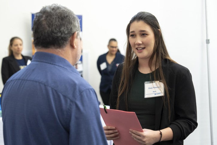 A woman in professional attire holds a pink folder while conversing with a man in a blue shirt.