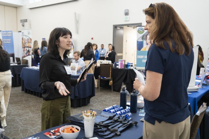 Two women converse at a busy career fair, one holding papers. Tables with promotional items and attendees networking fill the bright room. Vibrant, professional atmosphere.