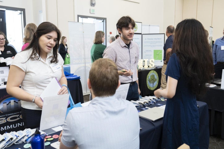 Two young adults engage in conversation with recruiters at a career fair booth. They appear attentive and engaged, surrounded by banners and informational materials.