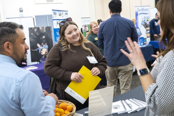 A woman with a yellow folder smiles while conversing with two recruiters. Other attendees and informational displays are visible.