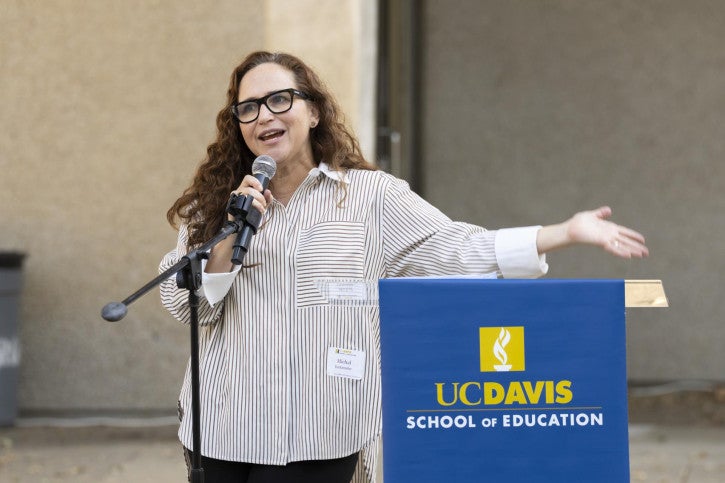Michal Kurlaender stands at a podium and addresses the event audience seated in front of her.