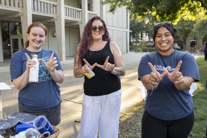 A PhD student, credential student, and staff member hold up their fingers in the shape of a "W," communicating that their team won one of the lawn games.