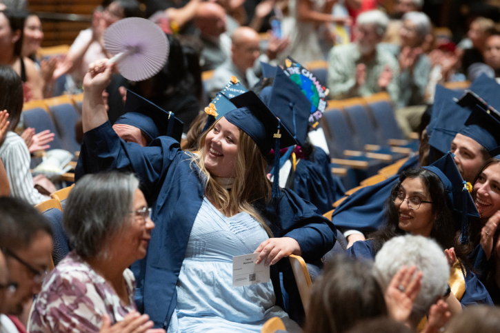 An MA grad smiles and waves to someone in the audience as she takes her seat.
