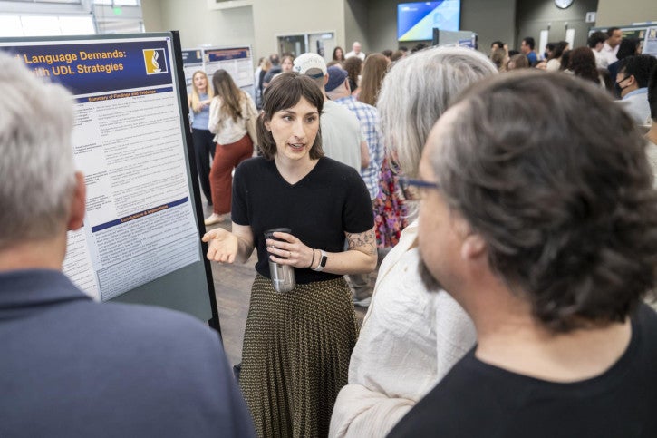 A woman in a black shirt and patterned skirt presents a research poster. She engages attentively with three attendees.