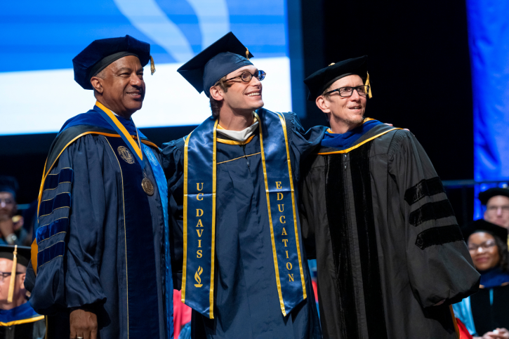 MA graduate Cameron smiles with Dean Smith and Chancellor May.