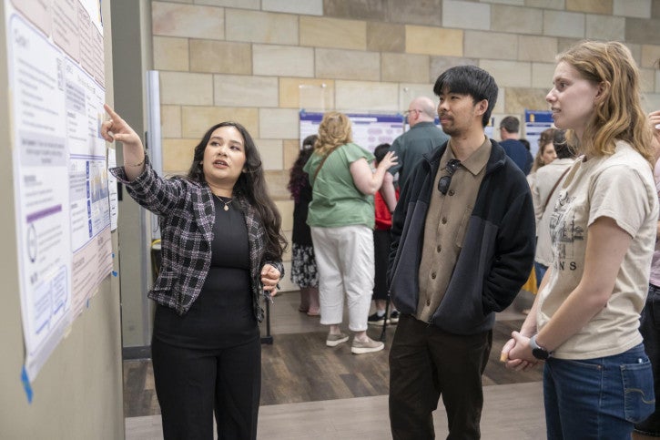 A woman in a plaid jacket presents a poster to a man and woman in casual attire. 