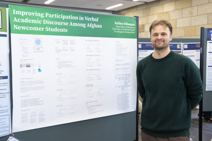 A man stands smiling beside a research poster titled "Improving Participation in Verbal Academic Discourse Among Afghan Newcomer Students." The poster includes charts and text.