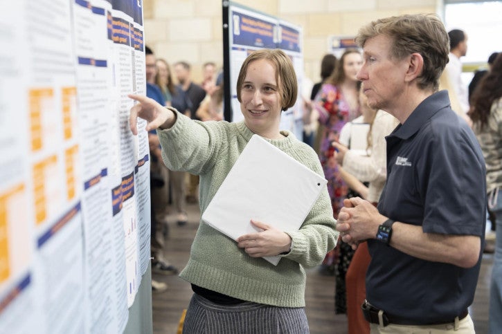 A woman with a folder explains a research poster to Dean Tom Smith. The atmosphere is engaged and academic, with others in the background.