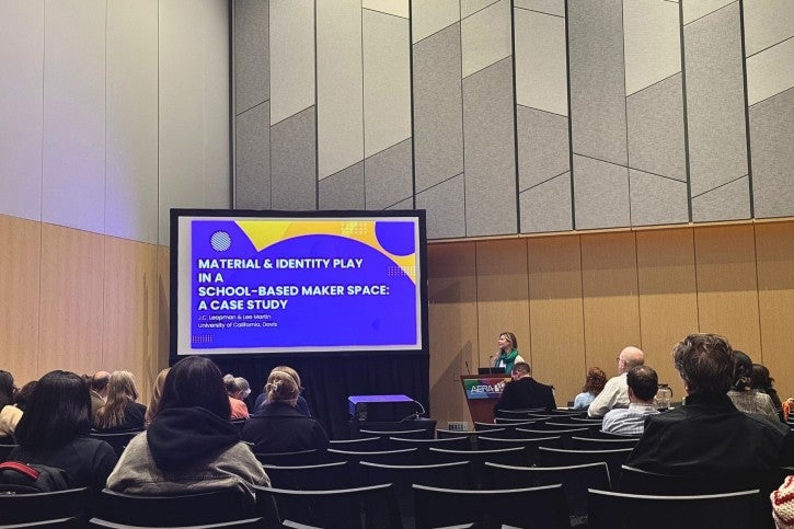 A person stands at a podium and speaks to an audience. Behind them, the presentation screen reads "Material and Identity Play in a School-Based Maker Space"