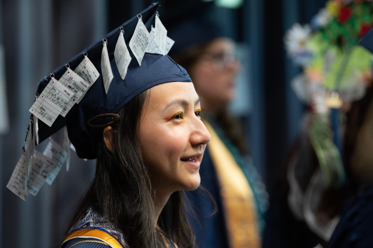 A credential student wearing a grad cap decorated with notes from their students smiles, looking into the distance.