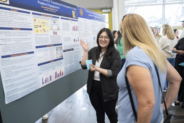 A woman enthusiastically presents research on posters with colorful graphs to an attendee. The setting is lively and engaged.