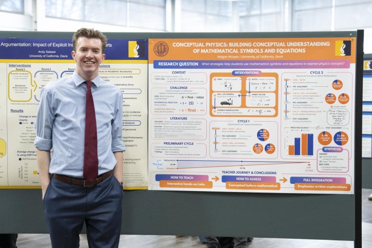 A man in a blue shirt and red tie stands smiling next to a colorful academic poster on physics education research.