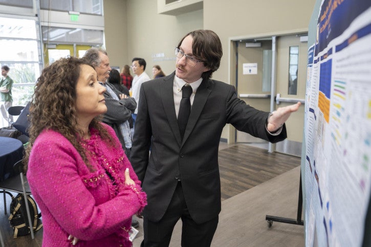 A man in a suit explains a poster presentation to a woman in a pink jacket, conveying focus and engagement. Other attendees are visible in the background.