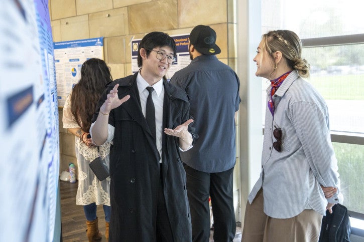 A man in a suit enthusiastically explains a poster to a smiling woman. 