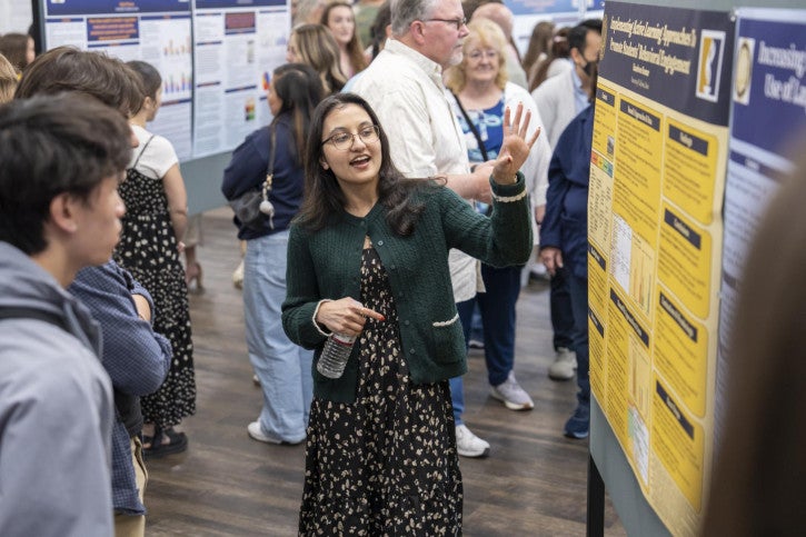 A woman in a green sweater enthusiastically presents a research poster to onlookers. The room buzzes with engaged attendees.