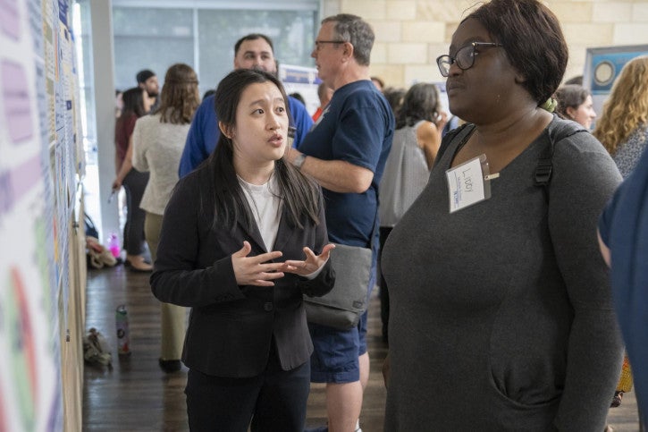 A woman in a blazer discusses a poster with an attentive attendee. The room is filled with engaged participants.