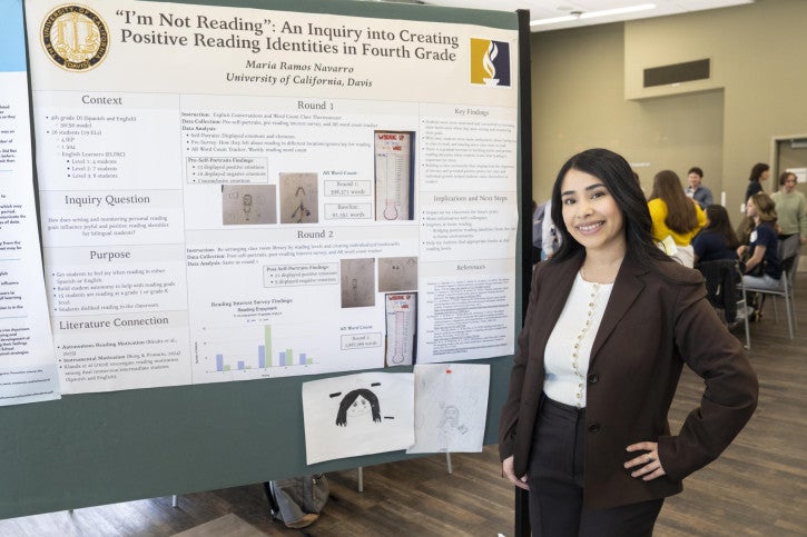 A smiling woman stands beside her academic poster titled “I'm Not Reading: An Inquiry into Creating Positive Reading Identities in Fourth Grade". People converse in the background.
