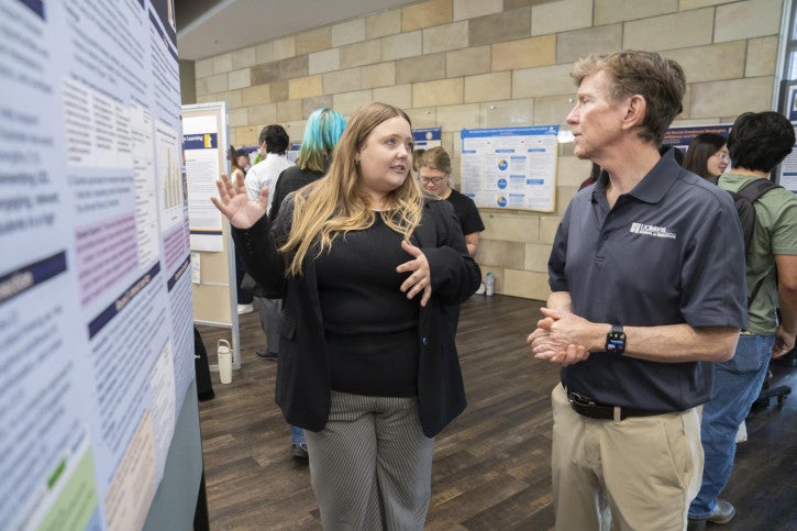 A woman enthusiastically explains a research poster to Dean Tom Smith. Other people discuss in the background.