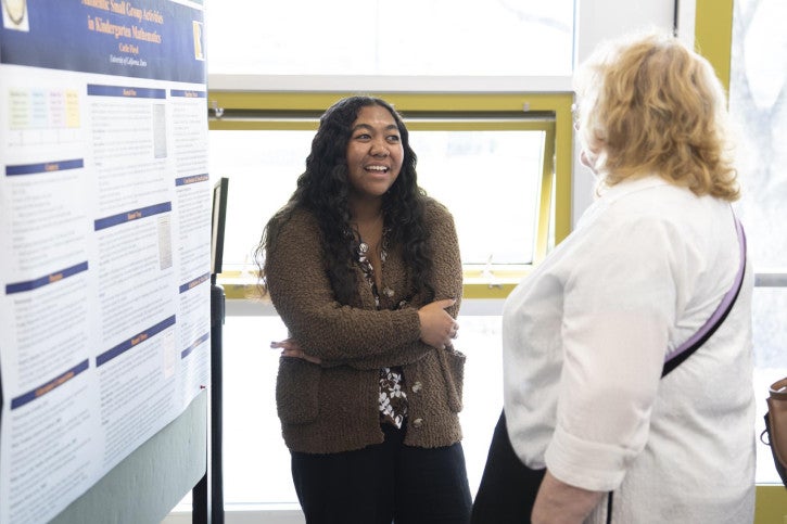 A woman with long, curly hair and a brown sweater smiles while discussing with another woman in front of a detailed research poster.