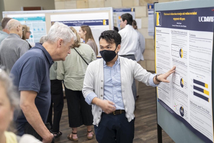 A person in a mask gestures at a research poster, explaining it to a man in a blue shirt. The atmosphere is focused and engaging.