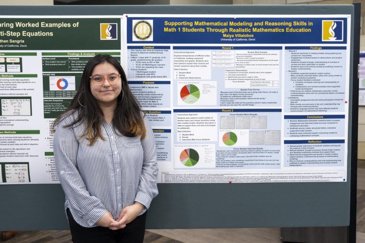 A woman wearing pink glasses stands smiling beside a large academic poster titled "Supporting Mathematical Modeling and Reasoning Skills in Math 1 Students Through Realistic Mathematics Education." The poster includes charts and text.