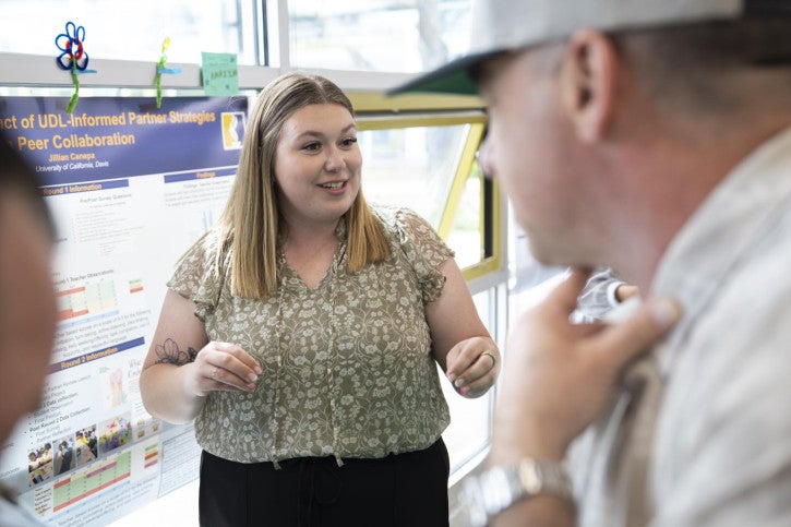 A woman presents a research poster on UDL-Informed Partner Strategies to a man, appearing engaged and enthusiastic in a bright, collaborative setting.