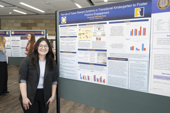 A person smiles next to a research poster on token reward systems in transitional kindergarten. The poster includes charts and colorful diagrams. Others are in the background viewing posters.
