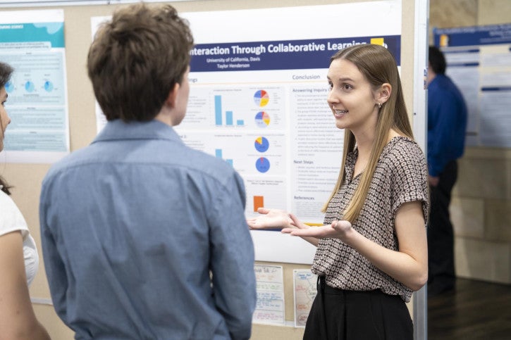 A woman enthusiastically explains a research poster to two attendees at a conference. The poster contains colorful graphs. The setting is professional and engaging.