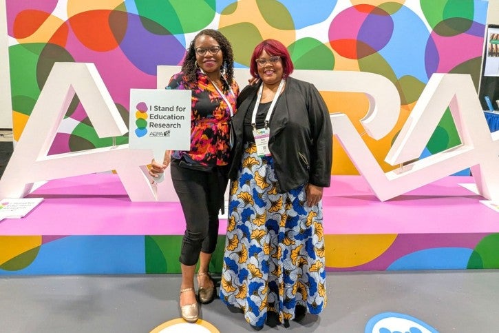Two women pose together in front of an oversized "AERA" display. The woman on the left holds a sign that reads "I stand for education research."