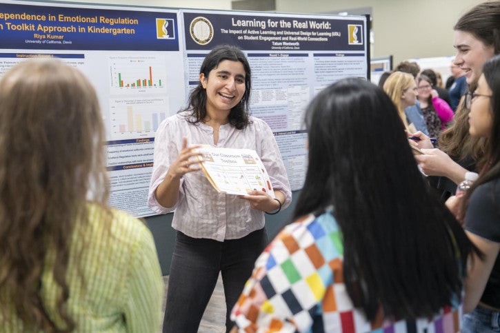 A young woman enthusiastically presents research at a poster session. She holds a document while talking to a small group. The atmosphere is engaging.