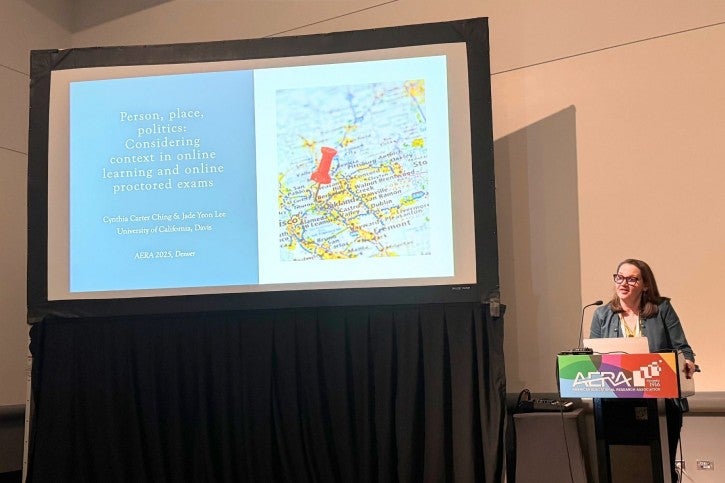 A woman stands at a podium and speaks into a microphone. Behind her, the presentation screen says "Person, Place, and Politics: Considering Context in Online Learning and Online Proctored Exams"
