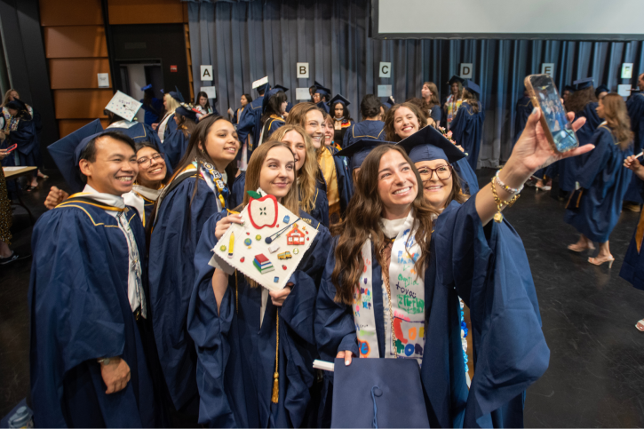 A group of MA students take a selfie. One holds up her grad cap, which is decorated with a felt apple, pencil, books, and schoolhouse.