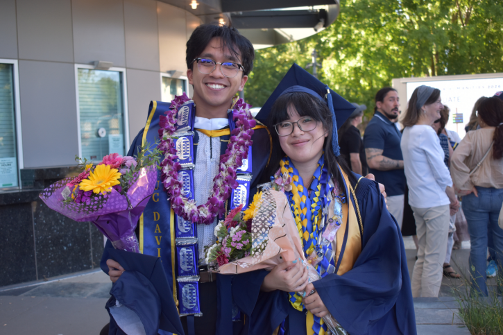 A credential and MA graduate stand together and smile, holding flowers and both wearing multiple leis.