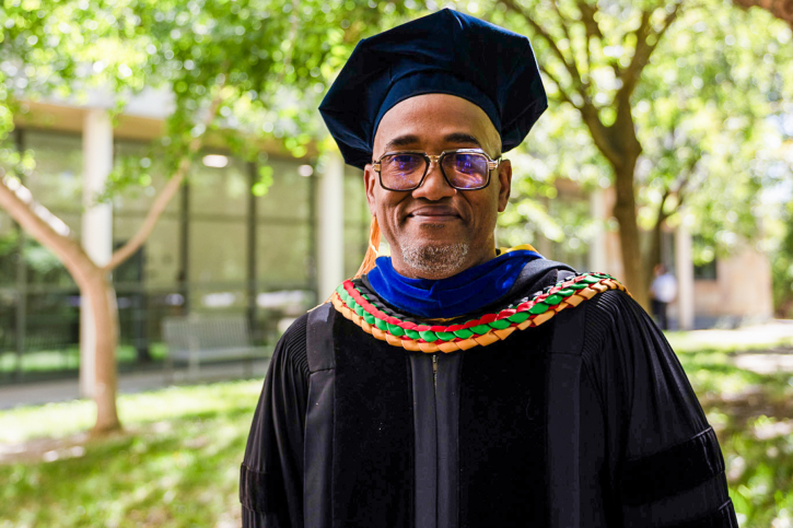 A PhD grad smiles for the camera, wearing regalia and a braided stole.