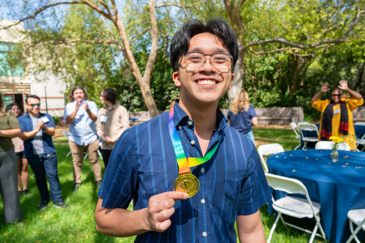 A student wearing glasses and a blue shirt holds up his gold medal.