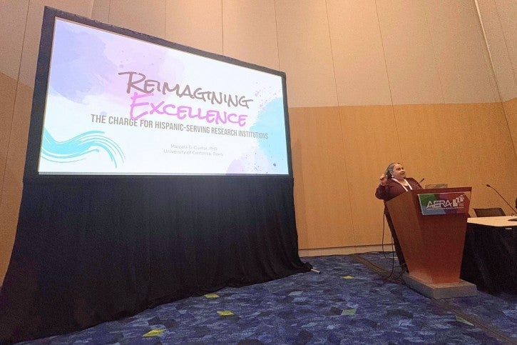 A woman stands at a podium and speaks into a microphone. Behind her, a presentation screen reads "Reimagining Excellence"
