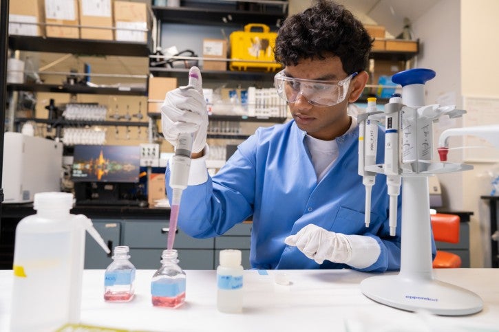 Student wearing gloves and blue lab coat works at lab station