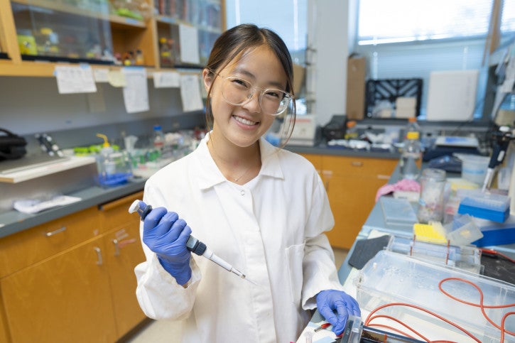 A student holds a pipette and smiles for the camera.