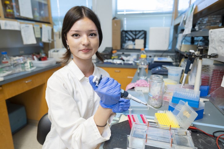 Ellen smiles for the camera, holding a pipette and sitting at the lab bench.