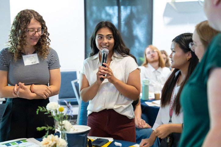 A group of five women stand up and address the room of people around them. One student speaks into a microphone.