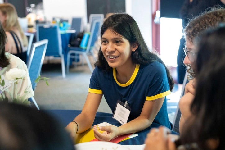 A credential student wearing a navy shirt leans over and smiles at people sitting in front of her.