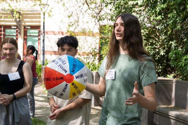 A student wearing a green shirt and nametag holds a beach ball in one hand and addresses a small group of people standing around them.